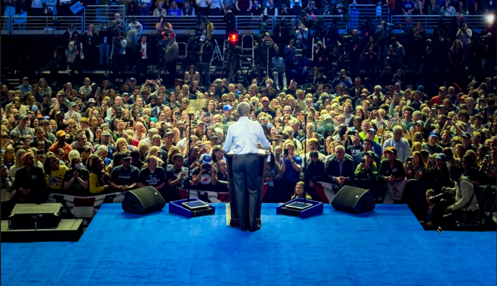 President Barack Obama speaks at a podium in front of a crowd. He stands on a blue stage. The image is show from directly behind him.
