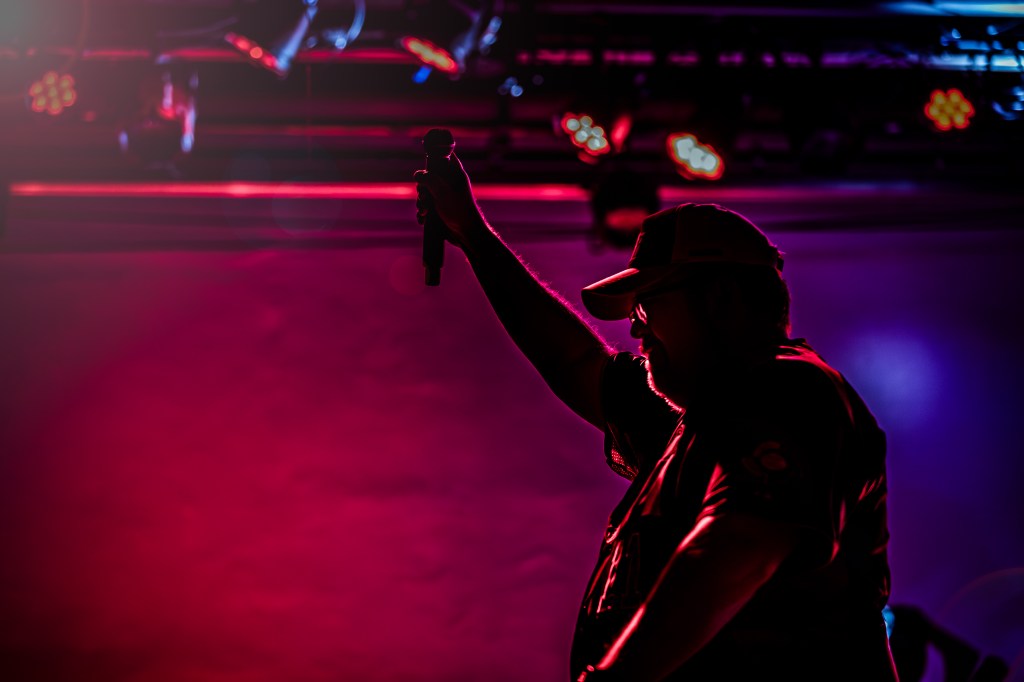 A pink and purple dark image of a singer on stage holding a microphone in an outstretched arm. He has a hat on.